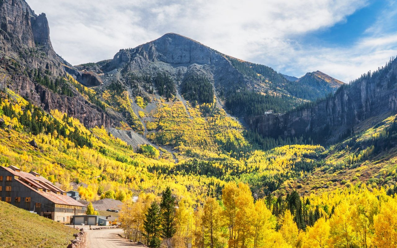 Golden aspens fill a rugged mountain valley around buildings at the foot of rocky peaks.