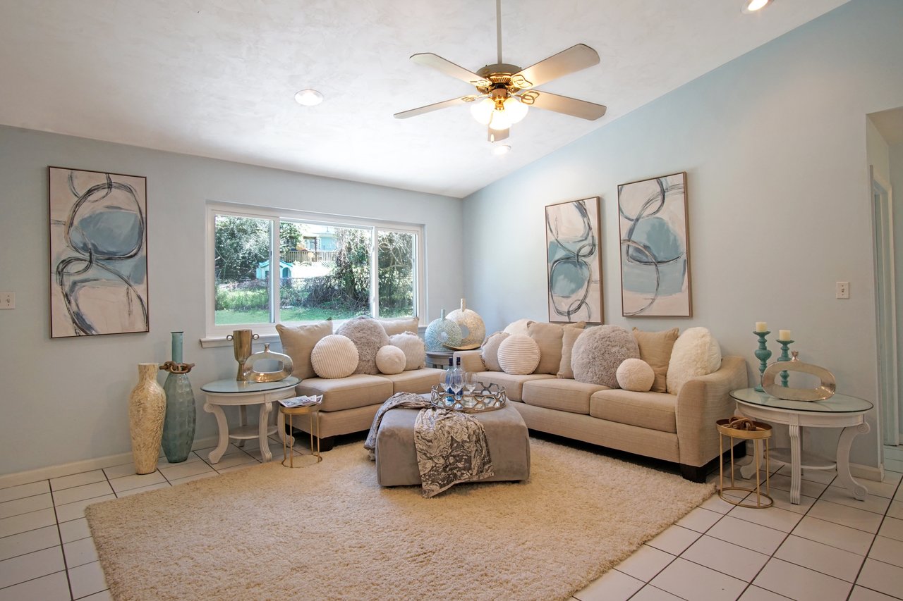 Bright living room with light blue walls, beige sofa, and round pillows. Modern abstract art, glass vases, and candles on side tables add elegance.