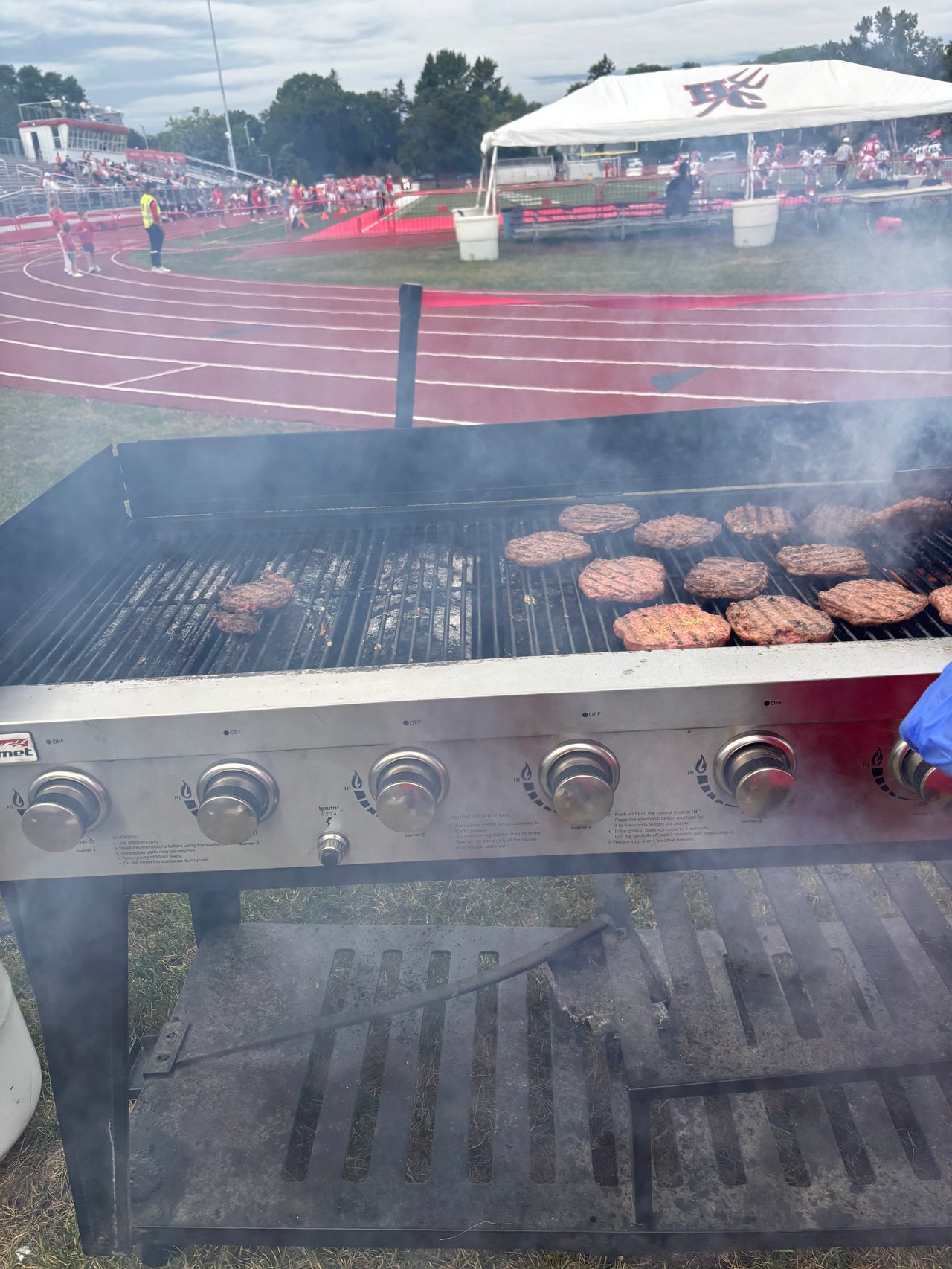 Burgers grill beside a high school football track, with players and a team tent beyond.