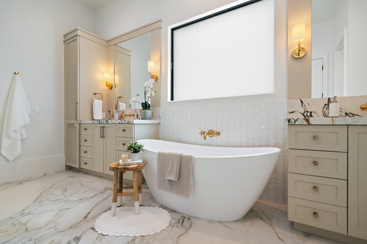 Bathroom with a freestanding soaking tub, gold fixtures, pale cabinetry and veined marble surfaces.