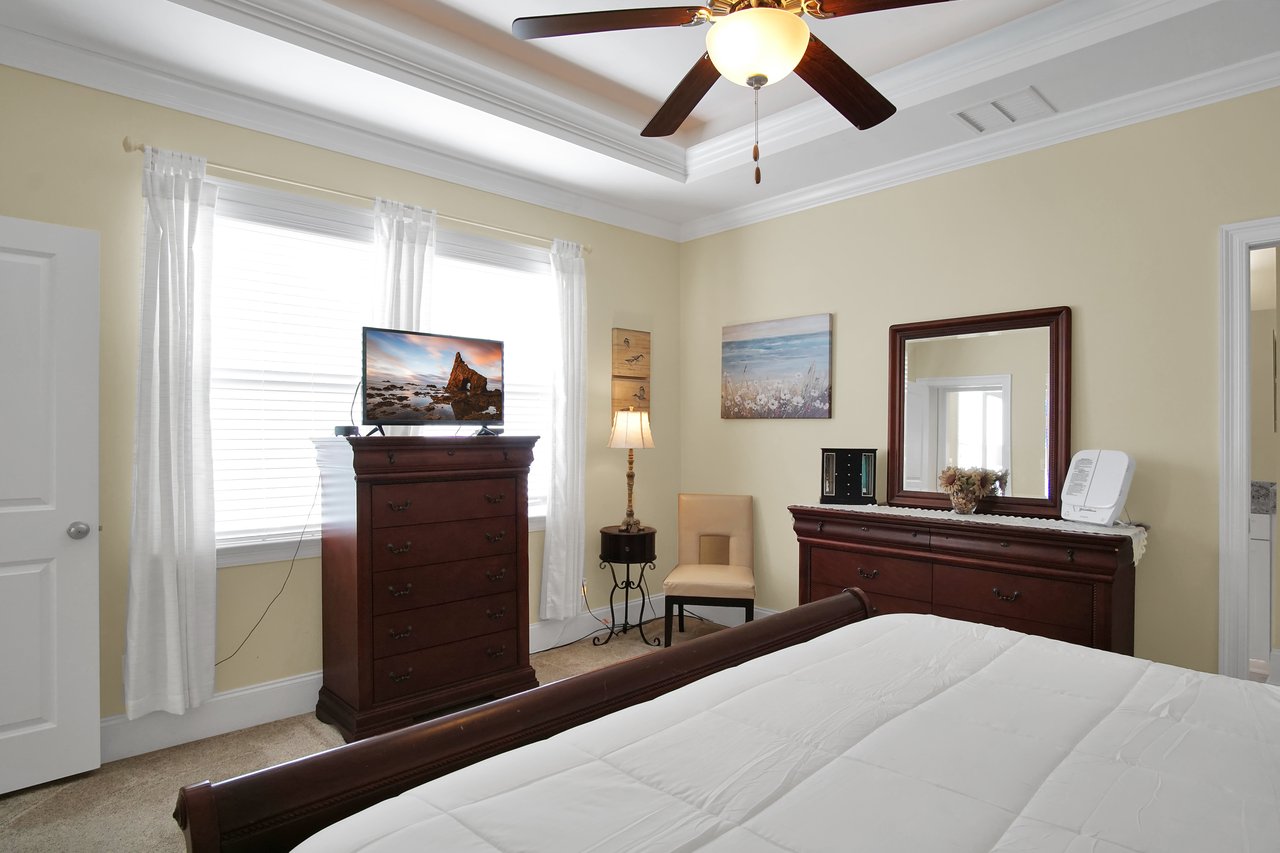 Bedroom with cream walls and large window, featuring wooden furniture, a ceiling fan, and a TV on the dresser. Warm and cozy ambiance.