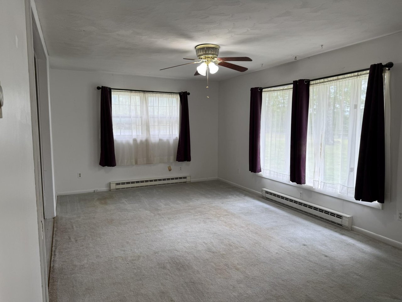 Empty living room with gray carpet, large curtained windows, and a ceiling fan.