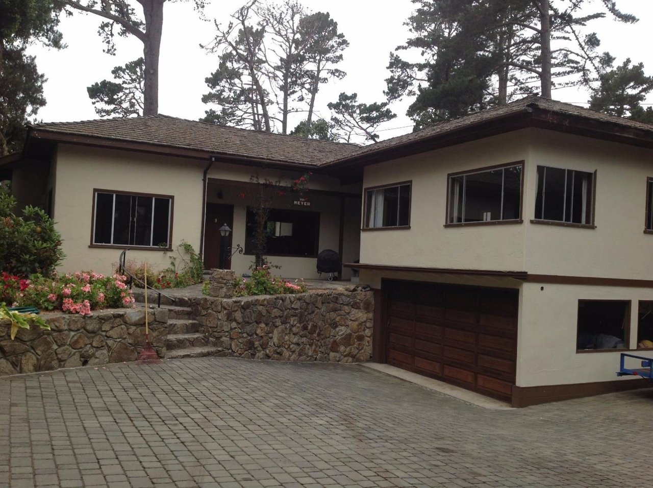 Split-level home with a paved driveway, stone retaining wall, and flowering garden beneath tall pines.