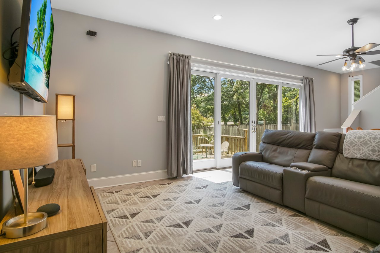 Bright living room with beige walls, a large gray sofa, and a geometric-patterned rug. Glass doors lead to greenery outside, creating a cozy, inviting space.