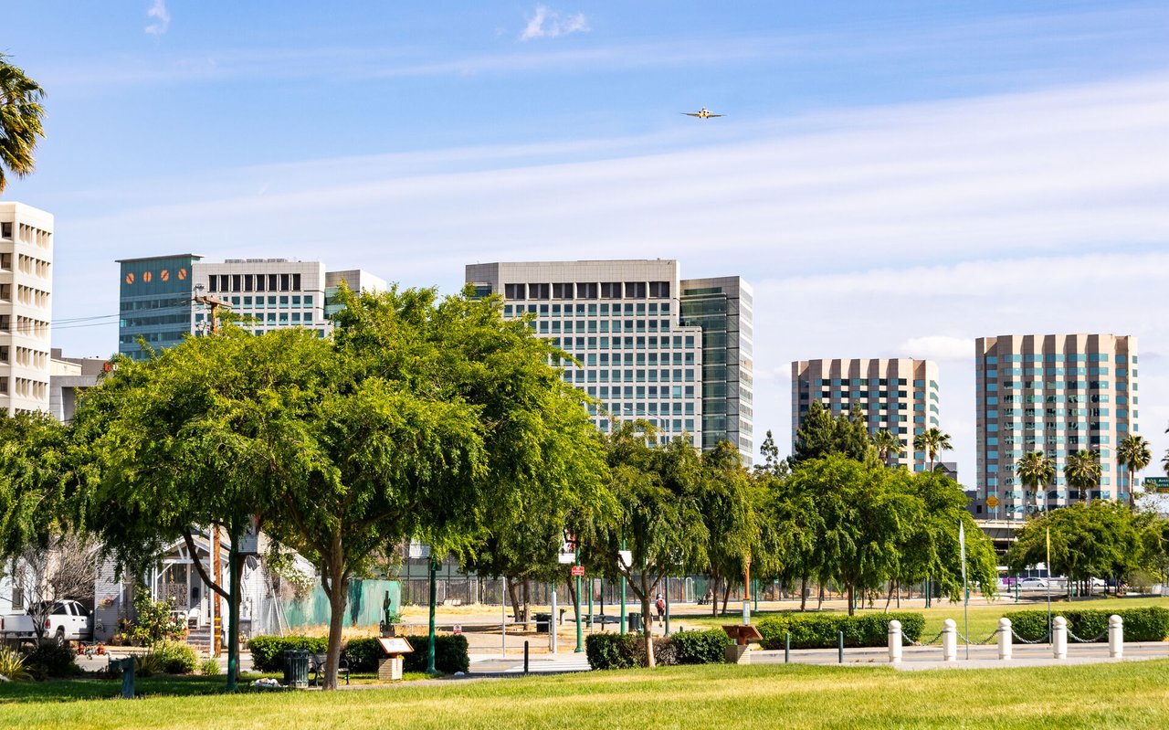 Downtown San Jose office buildings rise behind a tree-lined park beneath a passing airplane.