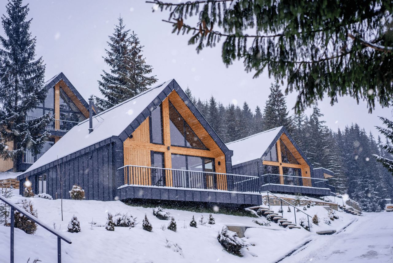 Modern A-frame homes with wood trim and snowy roofs among evergreen trees during a snowfall.