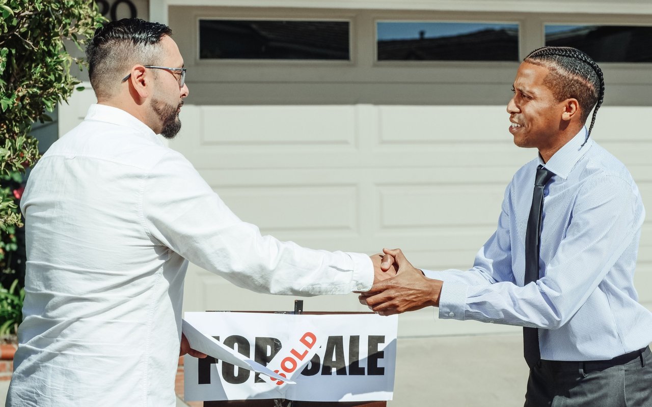 Two men shake hands beside a SOLD sign in front of a garage.