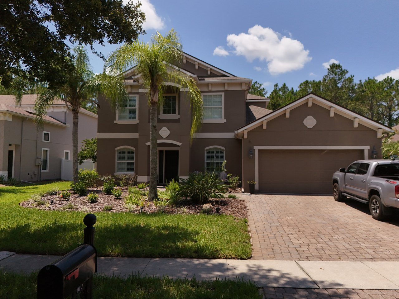 Two-story brown stucco home with a double garage, palm trees, and a pickup truck in the driveway.