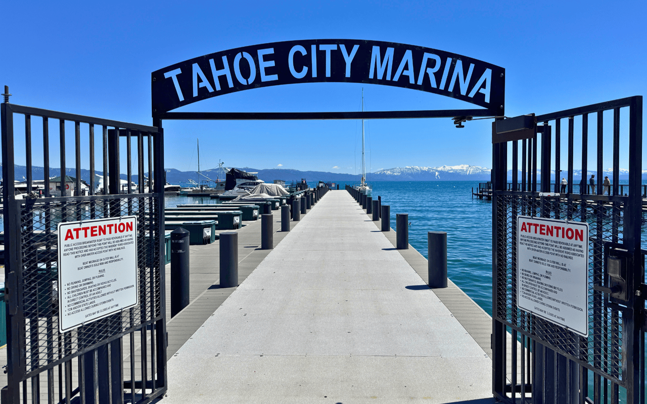 Tahoe City Marina’s entrance frames a dock extending into Lake Tahoe, with boats and snowy mountains beyond.