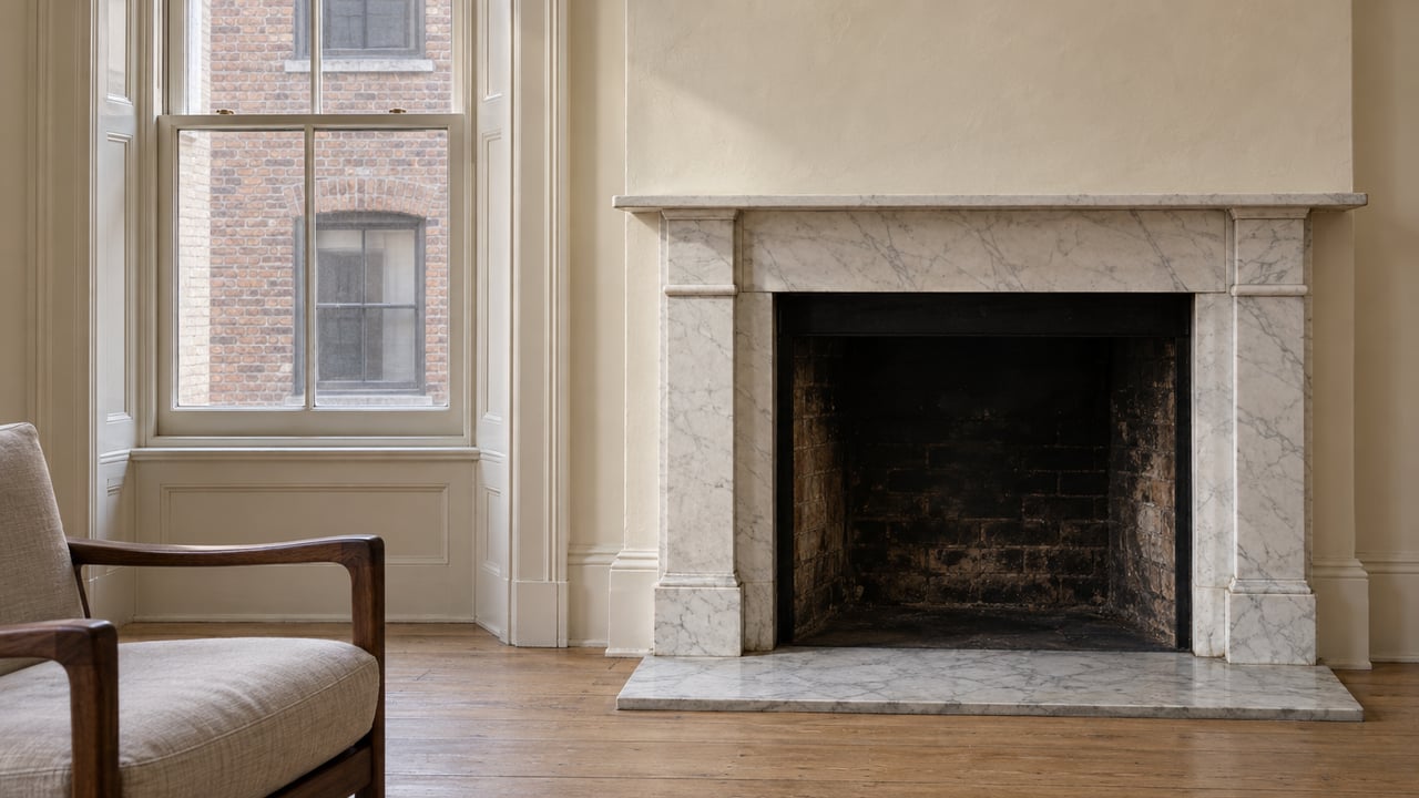 White marble fireplace beside a tall sash window, with pale oak flooring and the corner of a walnut and linen armchair.