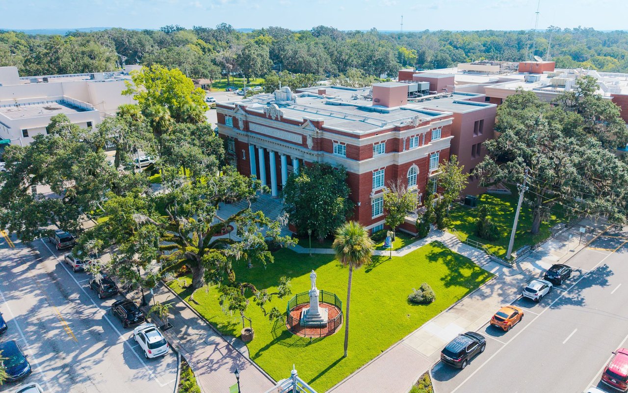 Aerial view of a red-brick civic building beside a tree-lined square and monument.