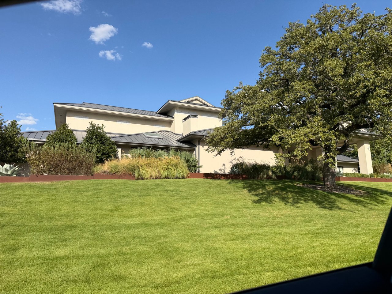 Modern cream-colored home with gray metal roofs, a broad lawn, and a large shade tree.