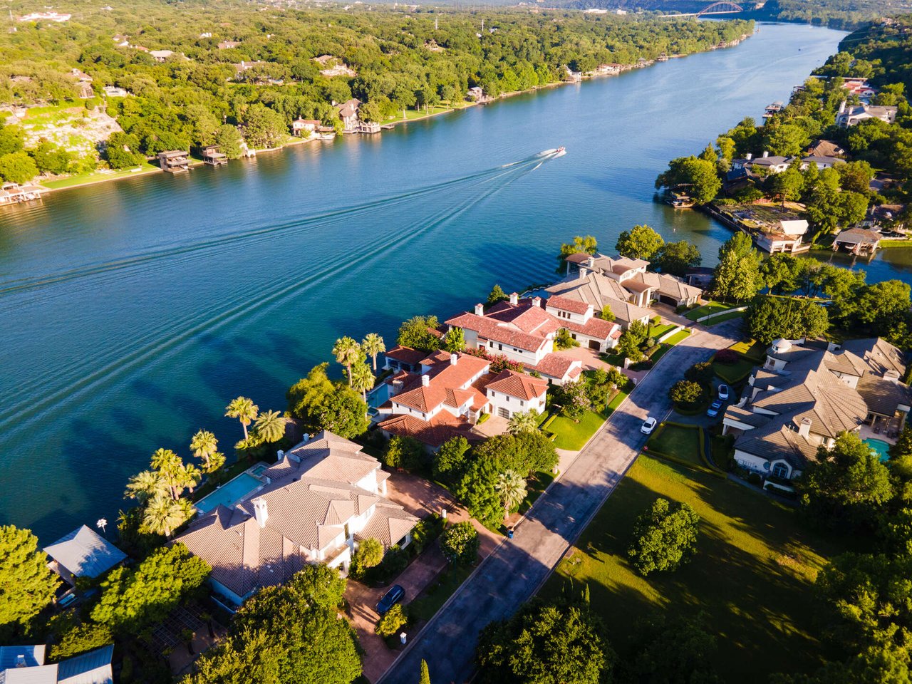 Aerial view of lakeside homes and tree-lined streets beside a boat-traced blue lake.