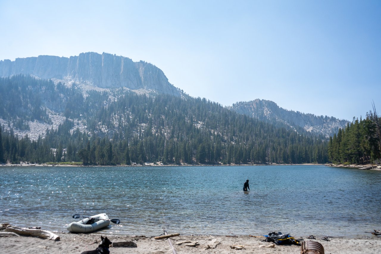 A person wades in mountain lake beneath forested granite peaks; inflatable raft rests on shore.