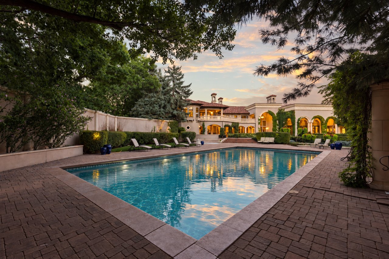 Estate pool and stone patio framed by mature trees, with the illuminated home beyond at dusk.