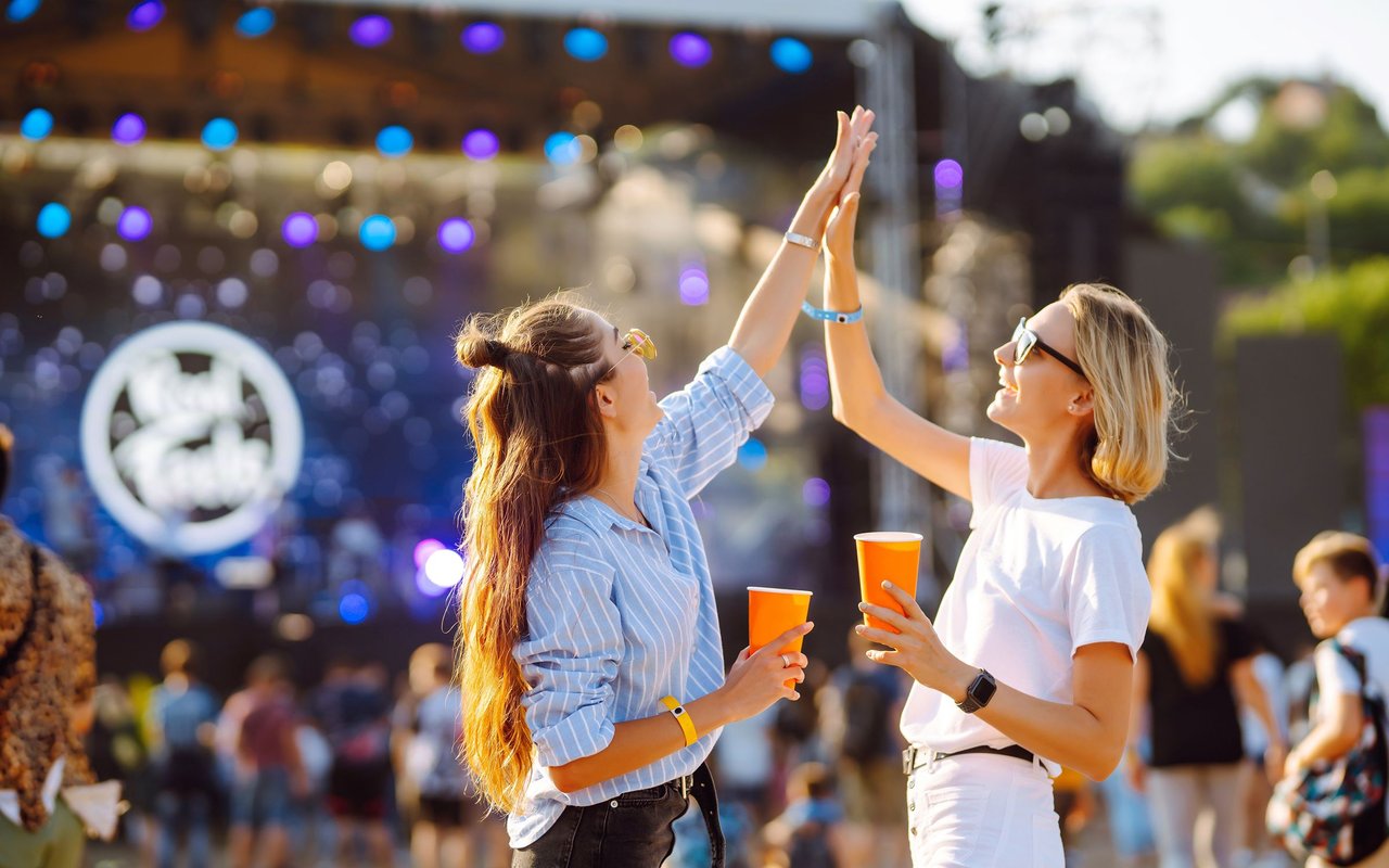 Two women high-five at a crowded outdoor event, holding orange cups beneath colorful stadium lights.