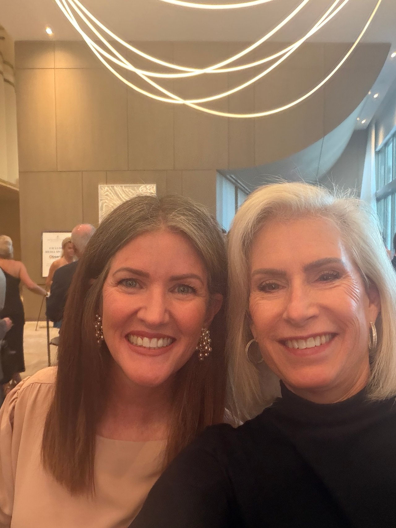 Two women smile for a selfie at an indoor event beneath looping ceiling lights.