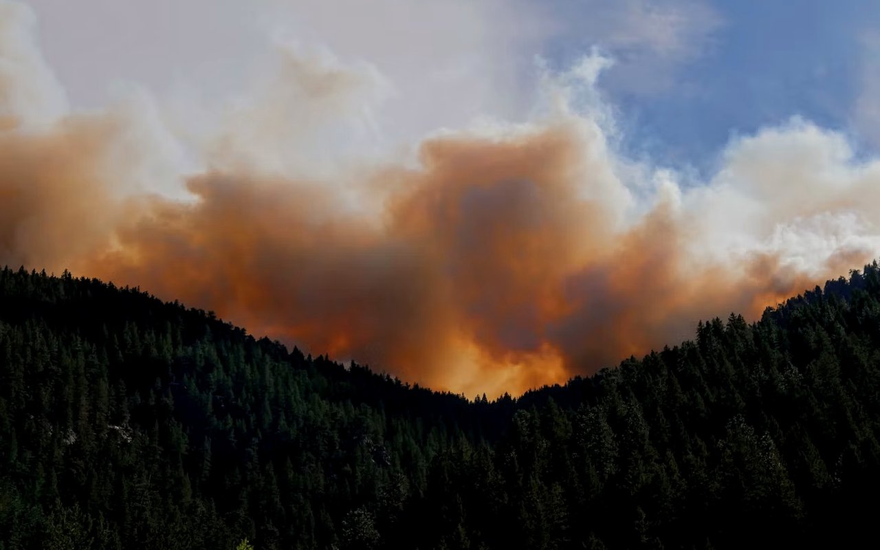 Orange-brown smoke billows behind forested mountain ridges under a partly cloudy sky.