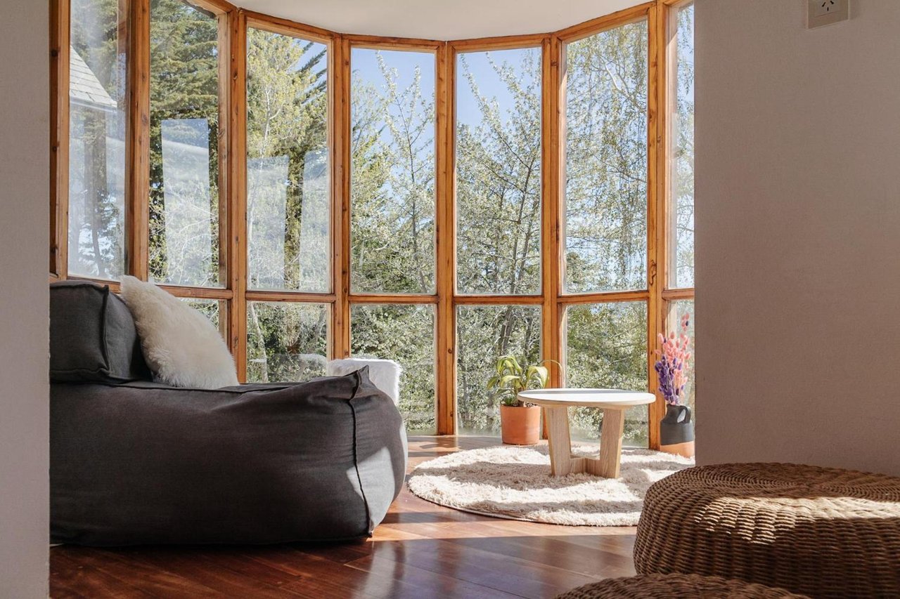 Living area with a dark sofa beside tall wood-framed windows overlooking trees.