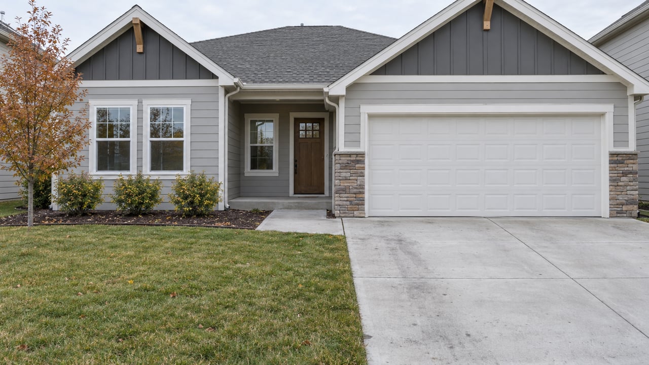Gray gabled home with an attached two-car garage, concrete driveway, small front lawn and a copper-leaved tree under soft autumn daylight.