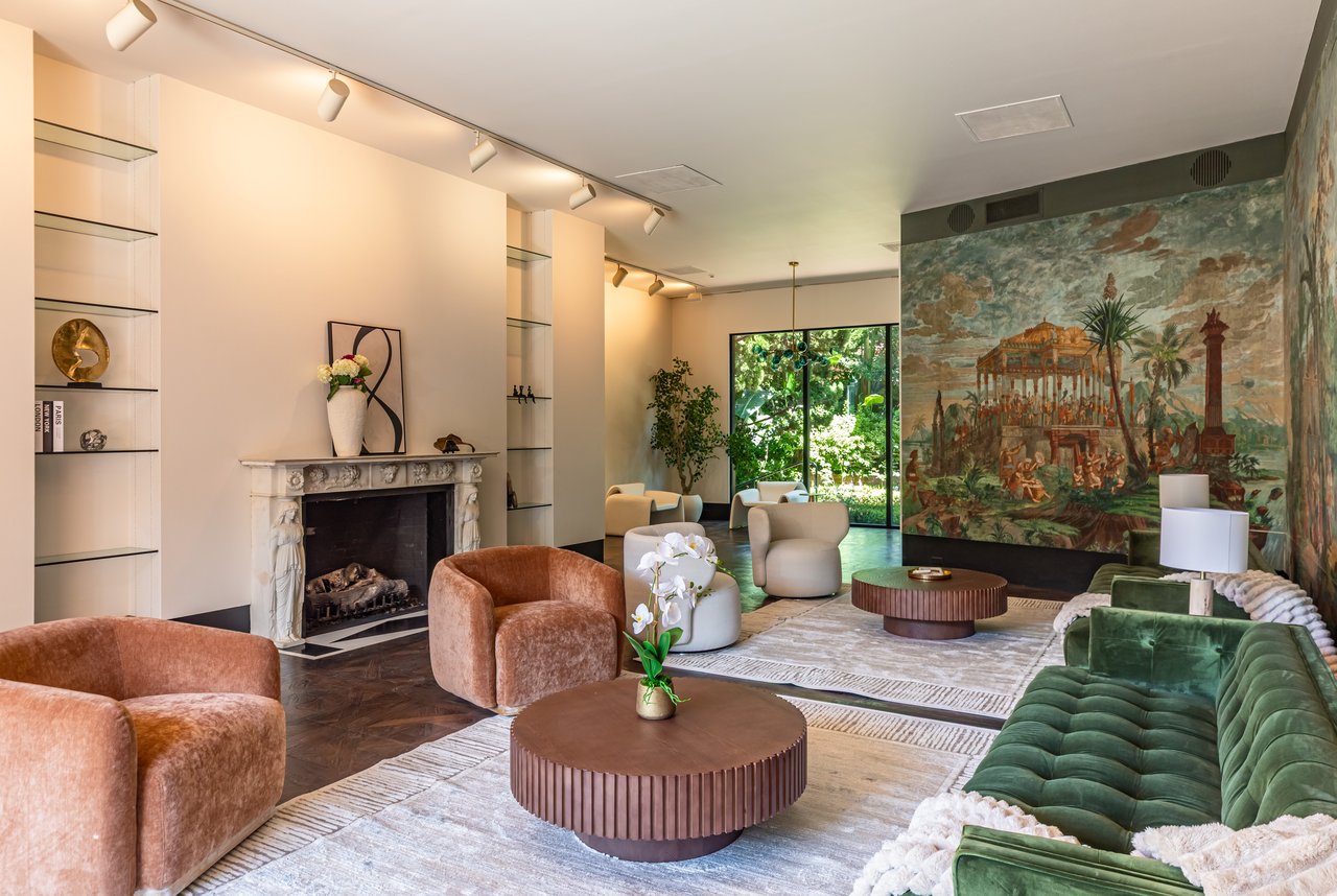 Living room with a carved fireplace, large mural, green velvet sofa, and garden views through glass doors.