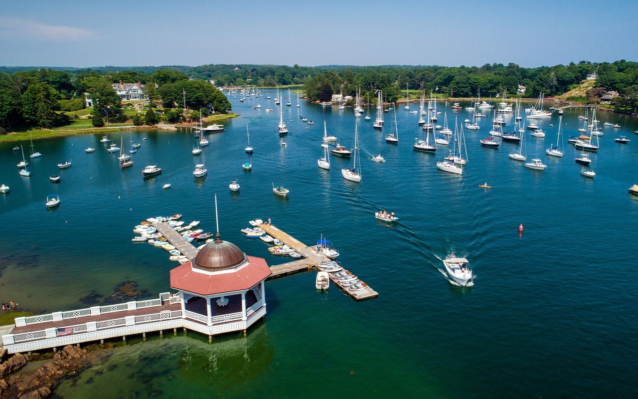 Aerial view of a wooded harbor filled with sailboats, with a waterfront pavilion in the foreground.