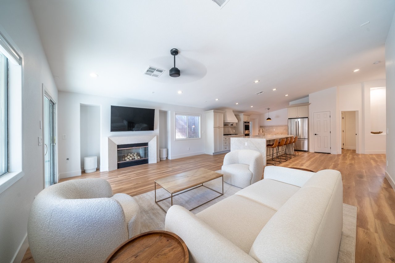 Open-plan living room with white seating, a fireplace and mounted TV, opening to a kitchen with an island.