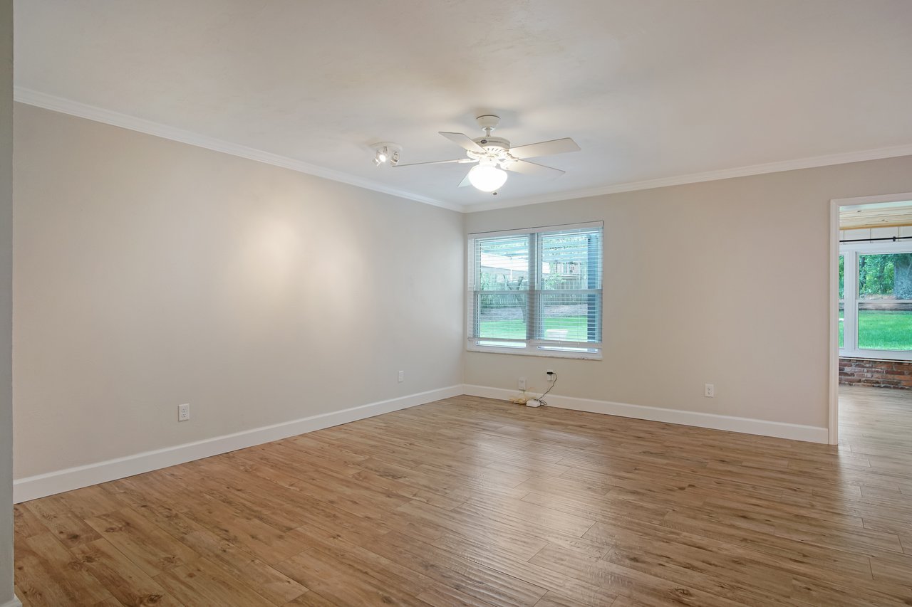 Spacious, empty room with light beige walls, wooden floors, and a ceiling fan. Large window on the right shows a green outdoor view, creating a serene ambiance.