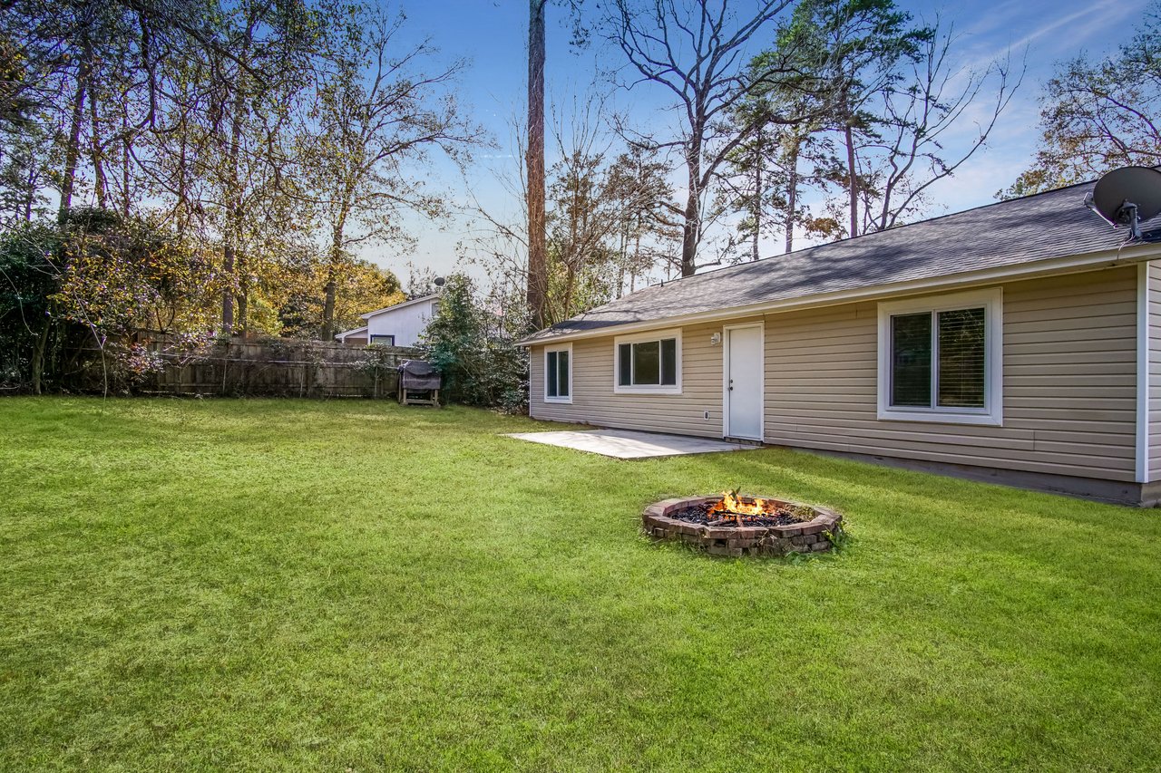 A backyard with green grass, a circular stone fire pit with a small flame, a beige house with large windows, and bare trees under a clear sky.