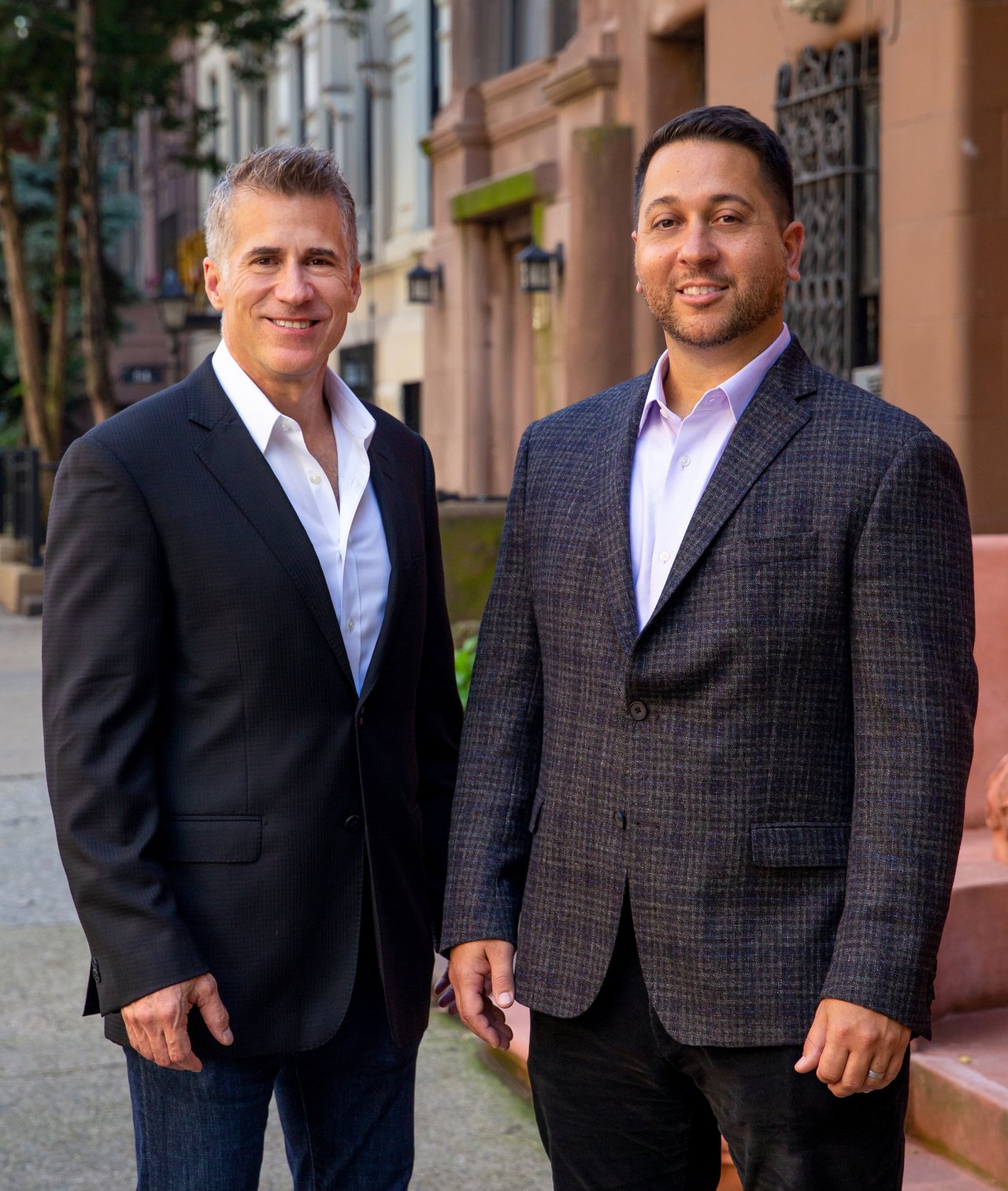 Two real estate agents in suits pose on a city sidewalk.