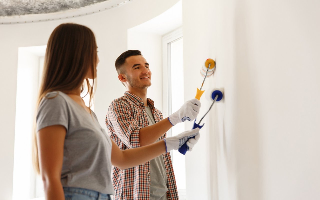 A couple paints a bright room together, using rollers on a white wall.