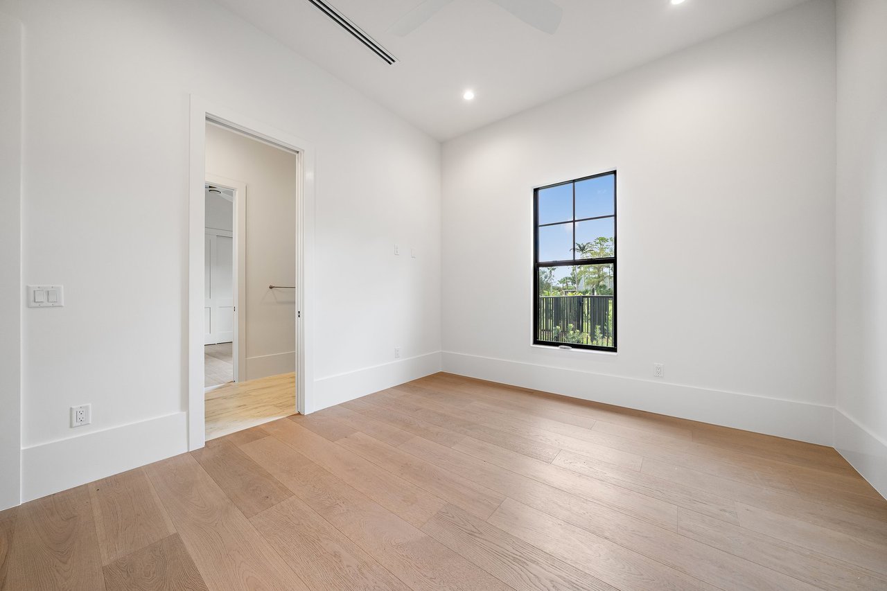 Empty bedroom with light wood floors, a tall black-framed window, and an open doorway.
