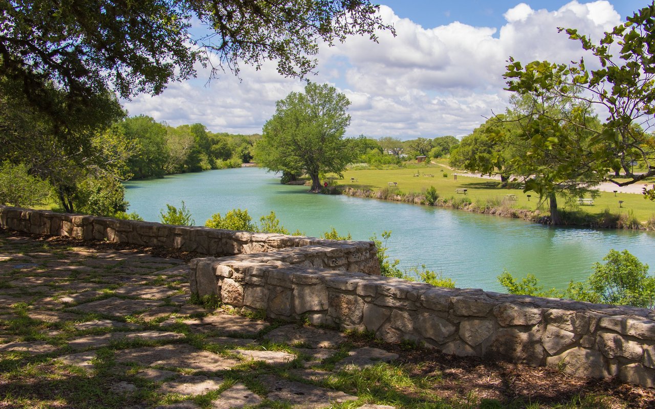 Tree-lined river and park viewed from a shaded path behind a low stone wall.