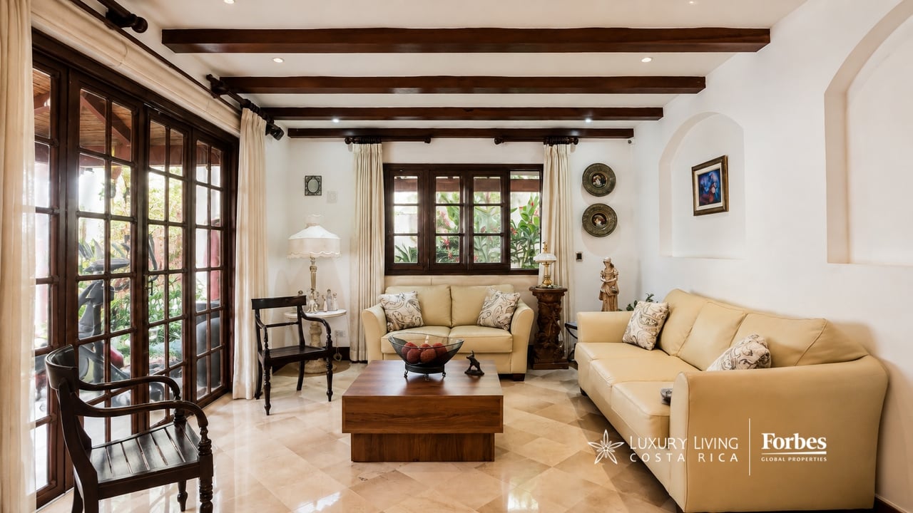 Living room with cream sofas, dark wood ceiling beams, and large windows overlooking greenery.