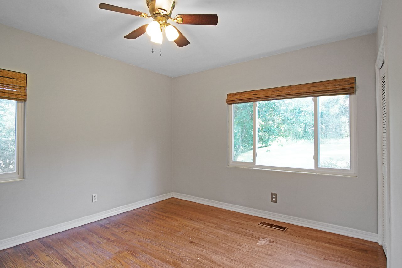 Empty room with beige walls, wooden floor, and ceiling fan. Large window with bamboo blinds lets in natural light, creating a calm, airy atmosphere.