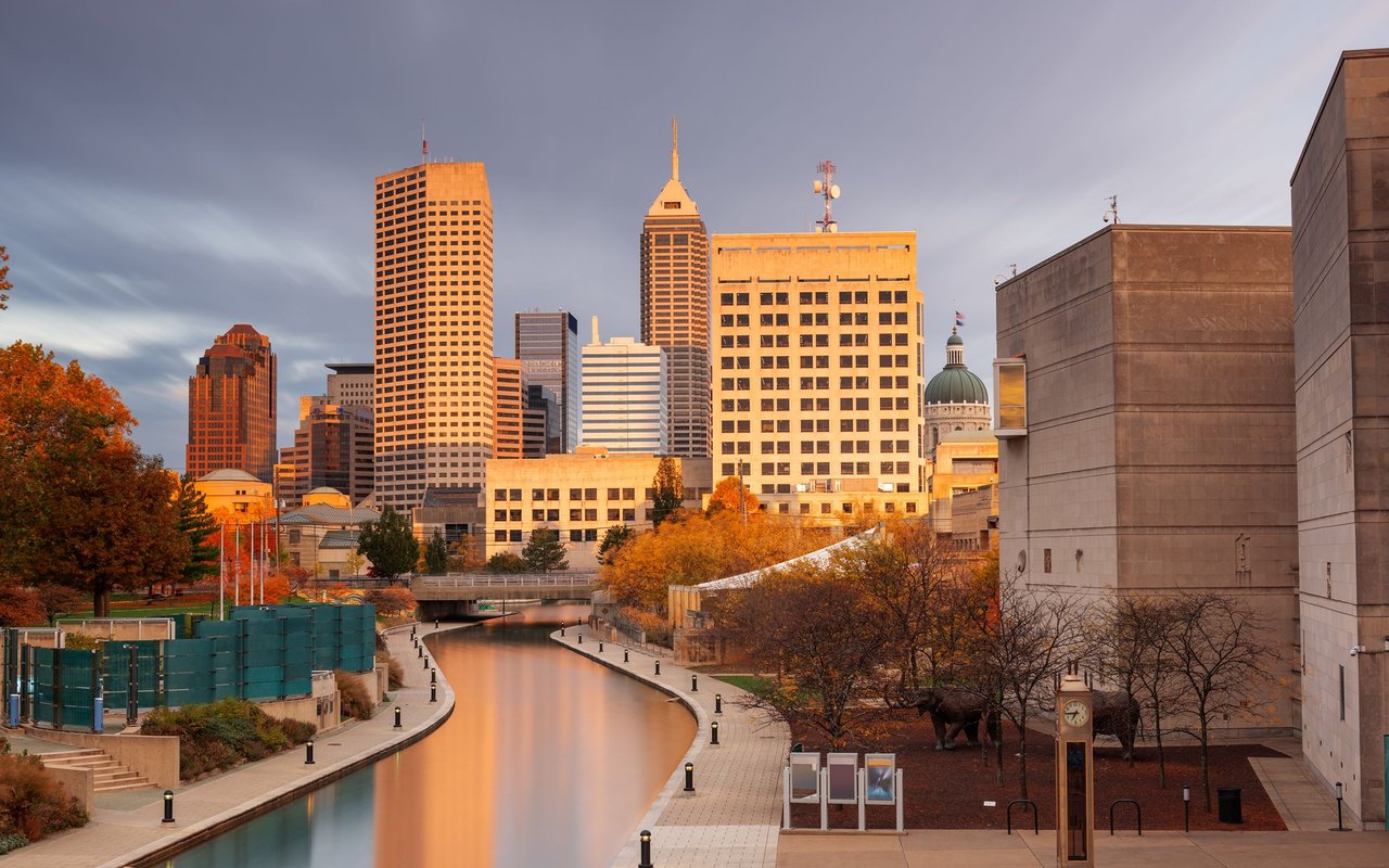 Downtown Indianapolis skyline and canal framed by autumn trees in golden-hour light.