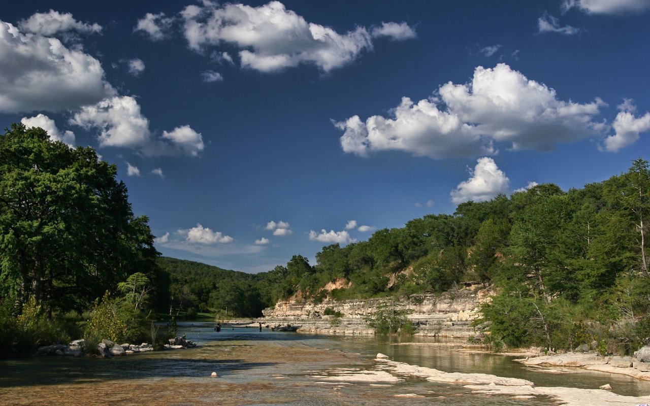 Shallow river over limestone shelves beneath wooded hills and a cloud-filled blue sky.