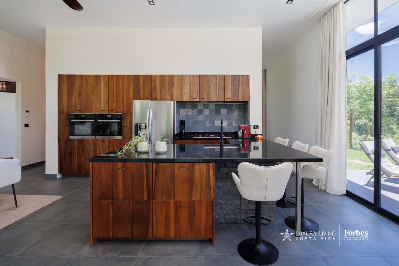 Modern kitchen with warm wood cabinetry, a large stone island, and floor-to-ceiling windows.