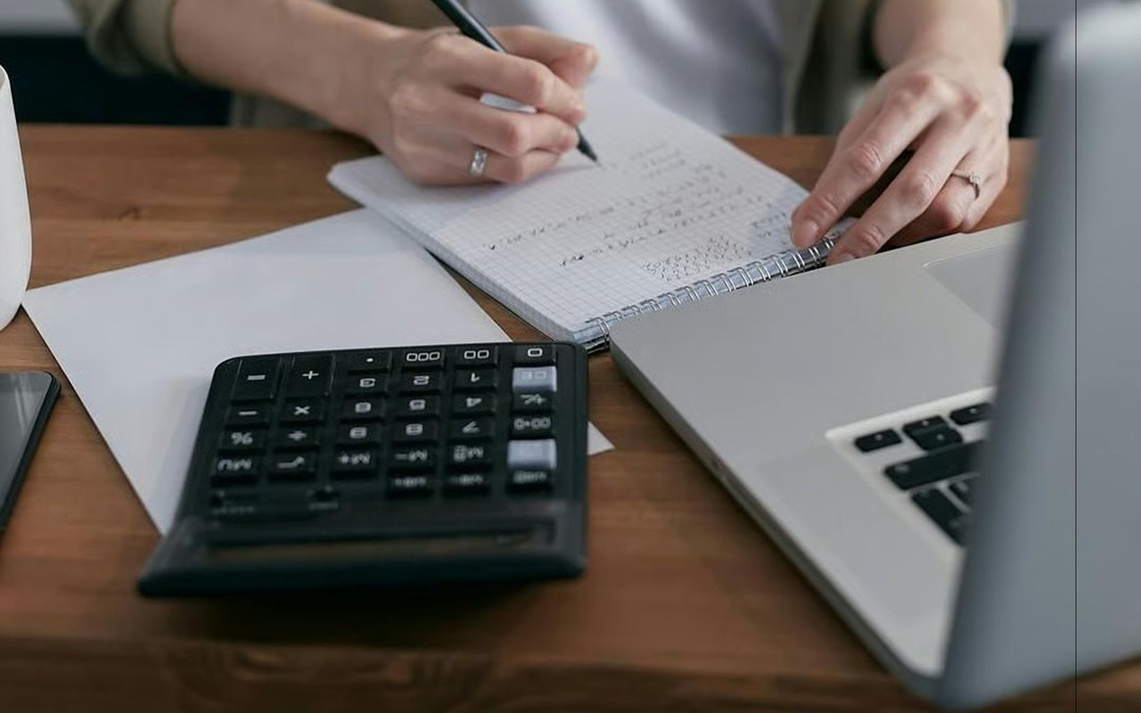 Hands writing in a notebook beside a calculator and laptop