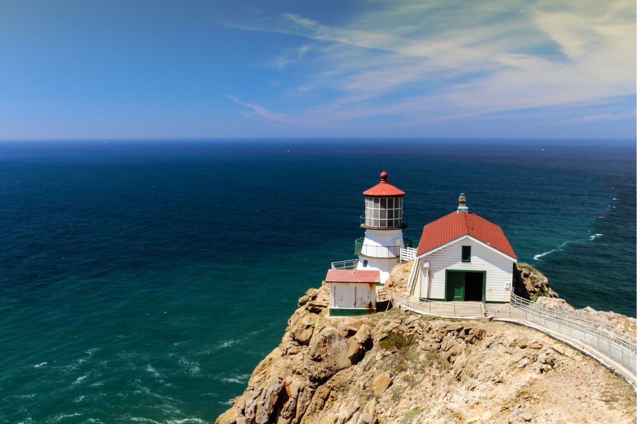 The Point Reyes Lighthouse, featuring a white tower and adjoining building with red roofs, perched on a steep, rocky cliff overlooking a vast blue ocean under a mostly clear sky.