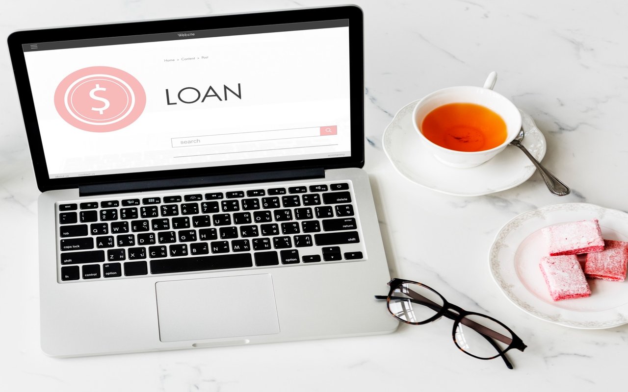 Laptop displaying a loan webpage beside tea, pink sweets, and eyeglasses on a marble table.