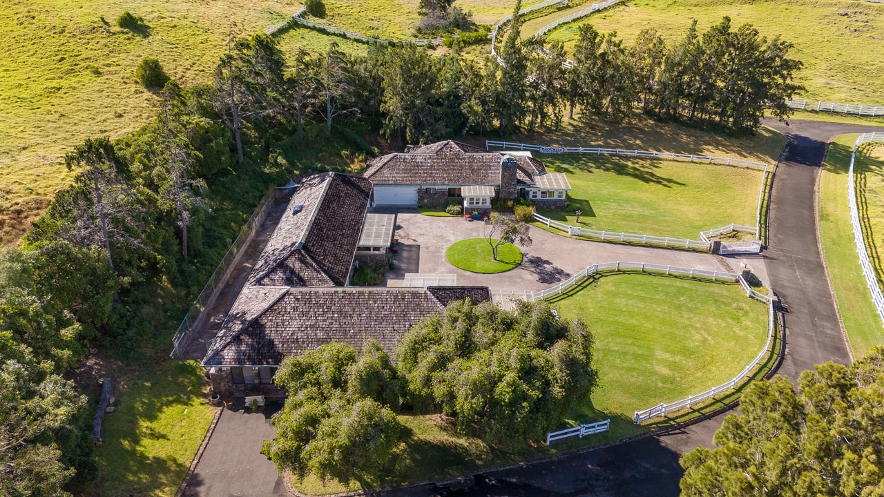 Aerial view of a large home with a circular driveway, fenced lawns, and surrounding trees.