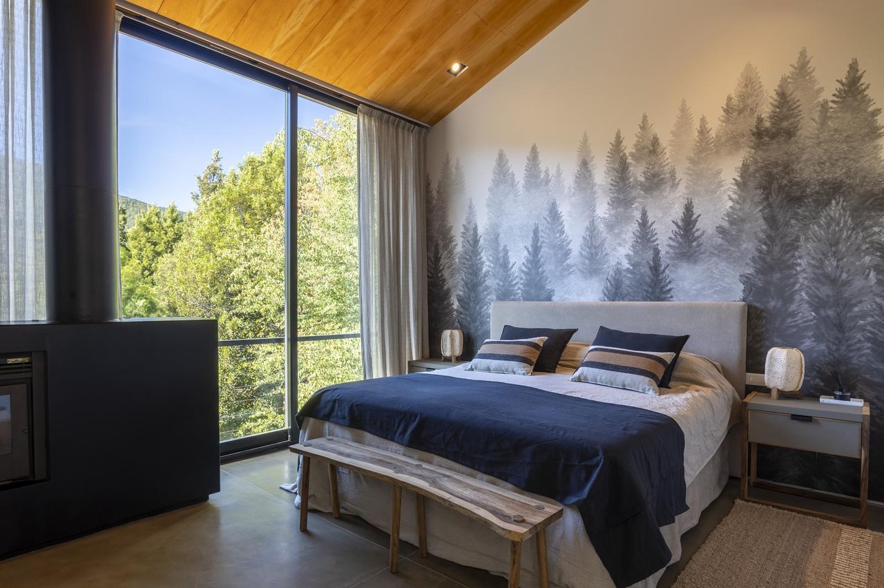 Bedroom with a forest mural, warm wood ceiling, and floor-to-ceiling windows overlooking trees.