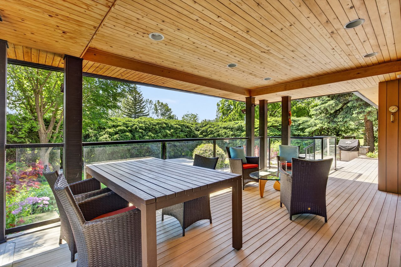 Covered wood deck with wicker dining set, glass railing, and views of a lush garden.