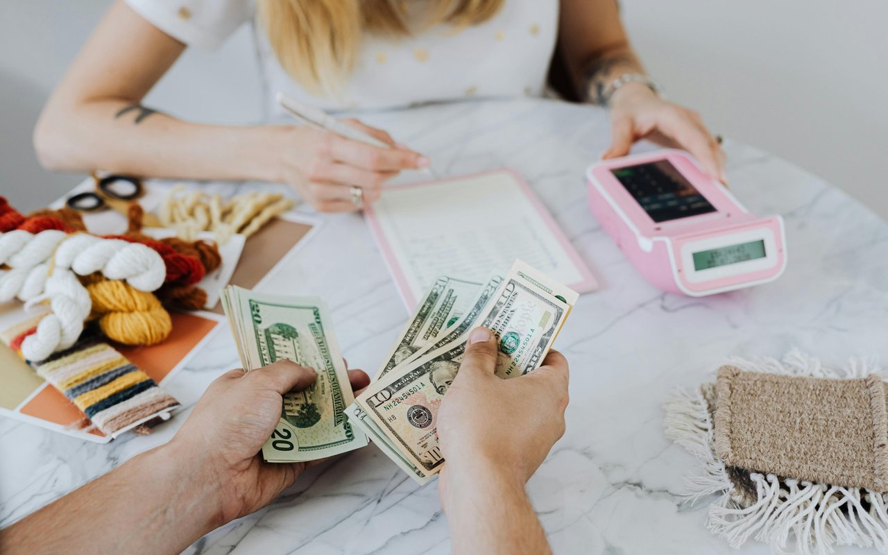 Hands counting U.S. cash beside a pink calculator and paperwork on a marble table.