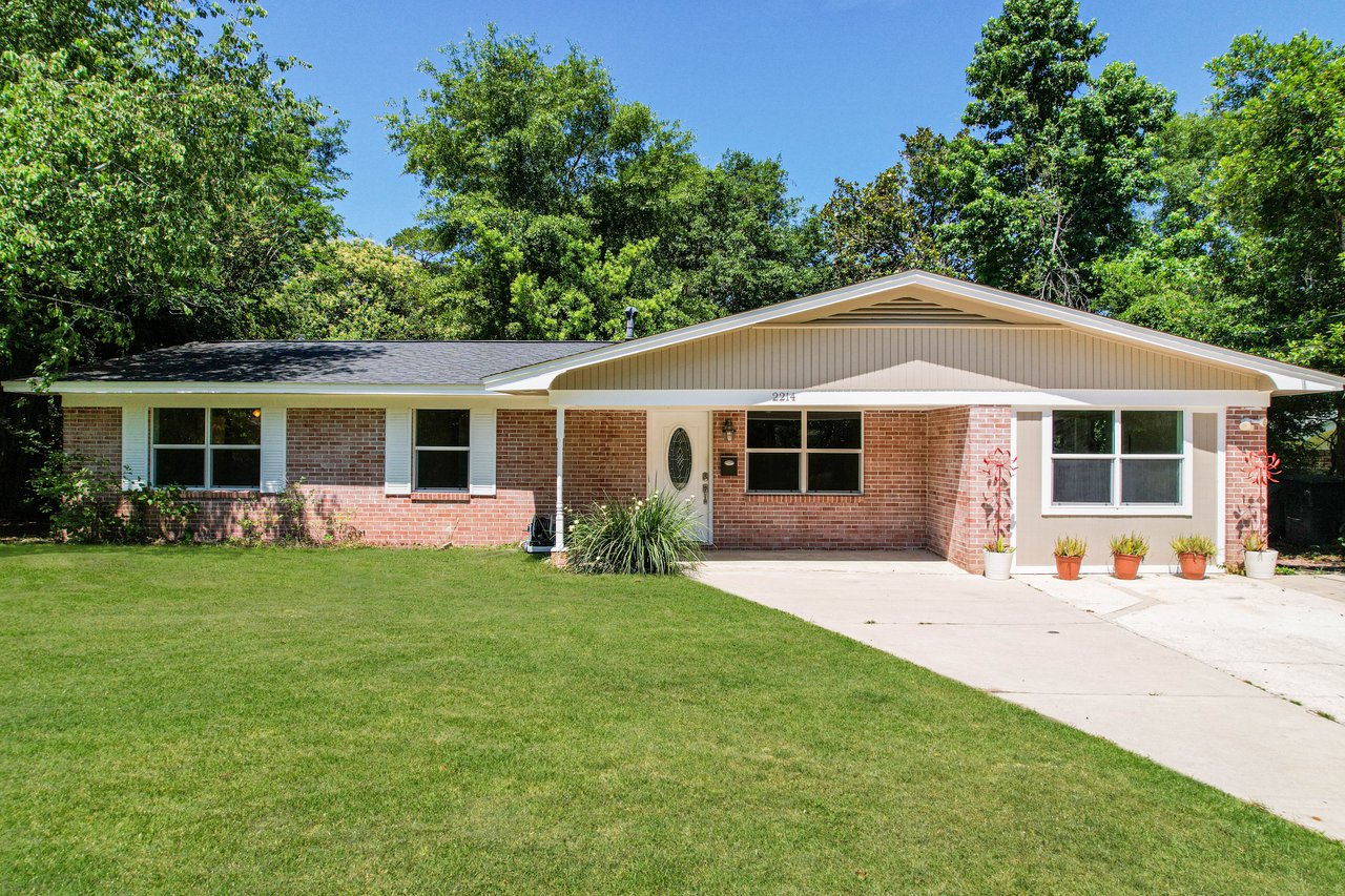 Single-story brick house with a wide driveway, large front lawn, and surrounded by lush green trees. Potted plants line the entrance, creating a welcoming look.