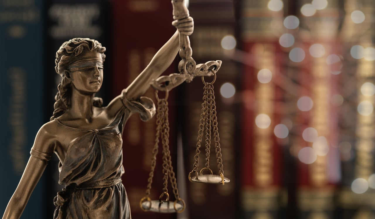 A Bronze Lady Justice statue stands blindfolded, holding scales, with law books blurred in the background. This iconic symbol of fairness and truth serves 