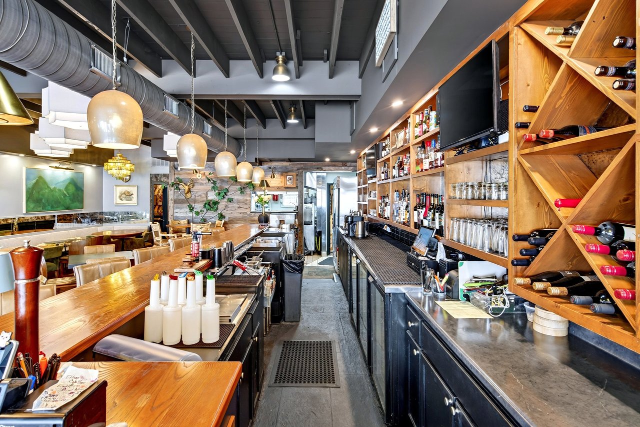 Restaurant interior with a long bar, wooden wine racks, dining tables, and exposed ceiling beams.
