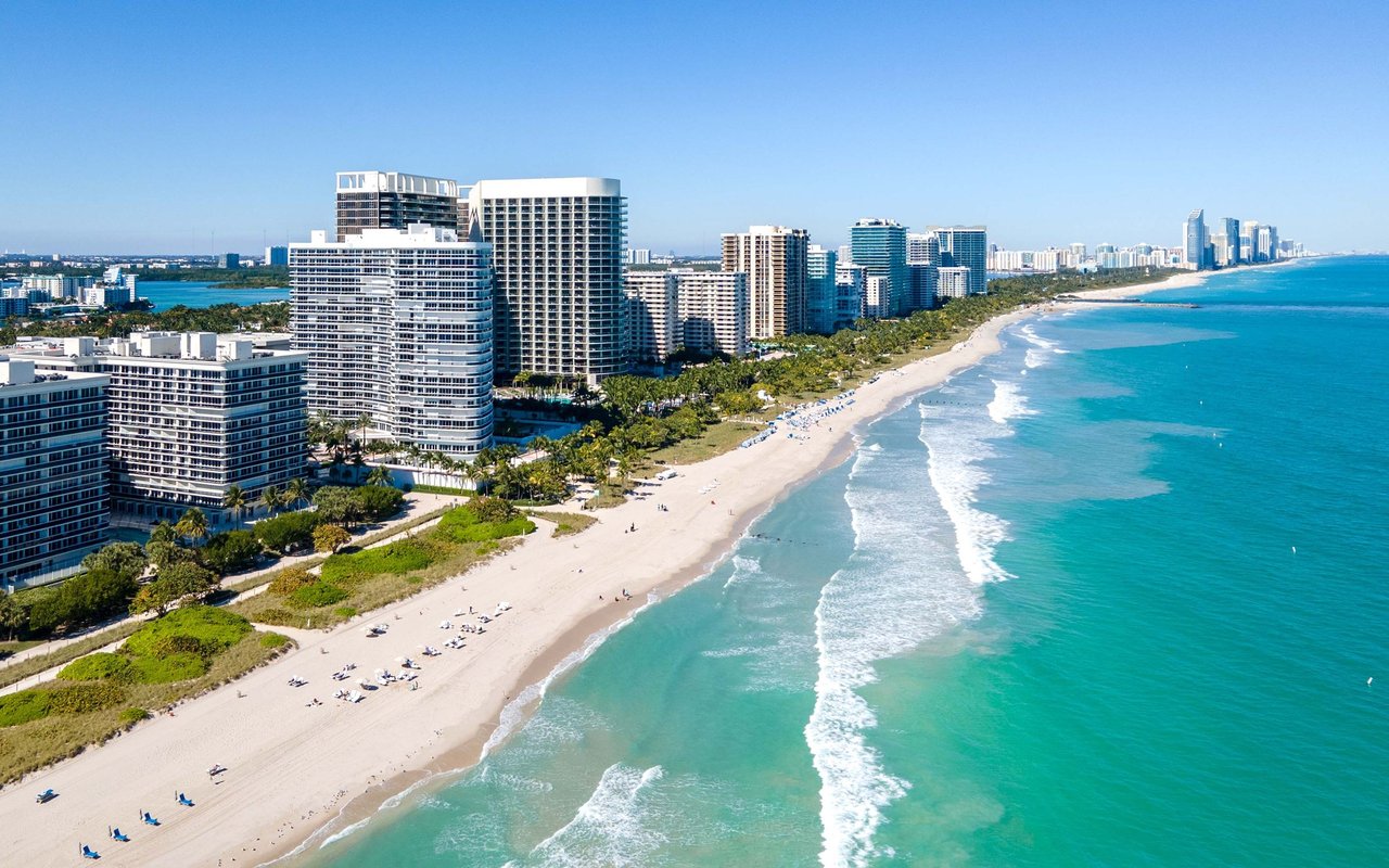 Aerial view of high-rise buildings lining a long sandy beach beside turquoise water.