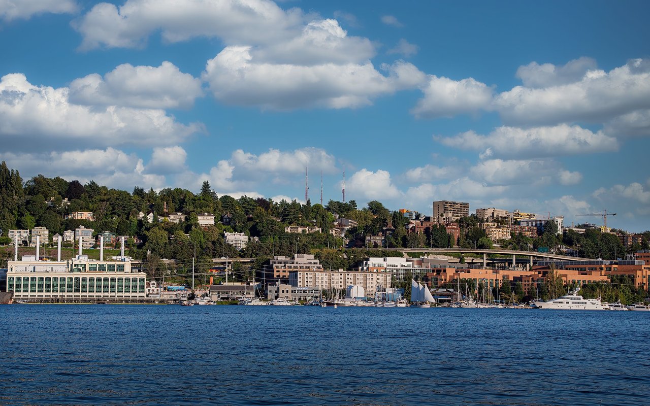 Waterfront marina with sailboats, shoreline buildings, and a wooded hillside under a cloudy blue sky.
